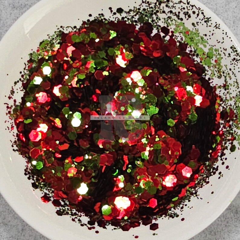 May include: Red, green and black glitter in a white bowl. The glitter is in a variety of shapes and sizes. "Island Glitters" is written in white on the glitter.