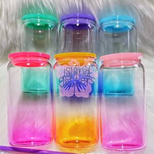 May include: A collection of clear glass jars with colorful lids and ombre designs. The jars have pink, orange, blue, green, and purple accents. One jar features the text "island glitters" with a floral design.