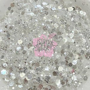 May include: Close-up of a pile of white and silver glitter. The glitter pieces are various shapes and sizes, including hexagonal and round. The image has a bright, reflective quality. The text "island glitters" is in the center.