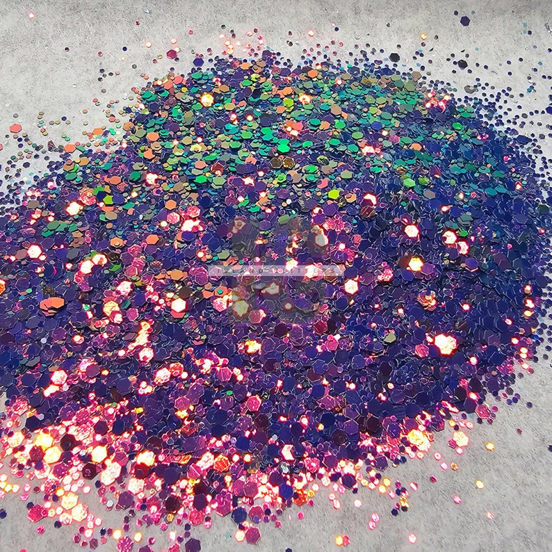 May include: A close-up of iridescent glitter in shades of purple, pink, and green. The glitter is scattered on a white surface.