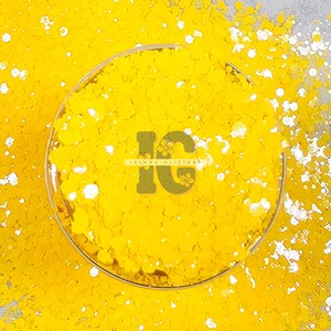 May include: A close-up of a yellow glitter with white glitter flakes. The glitter is in a clear glass jar with the text "Island Glitter" on the jar.
