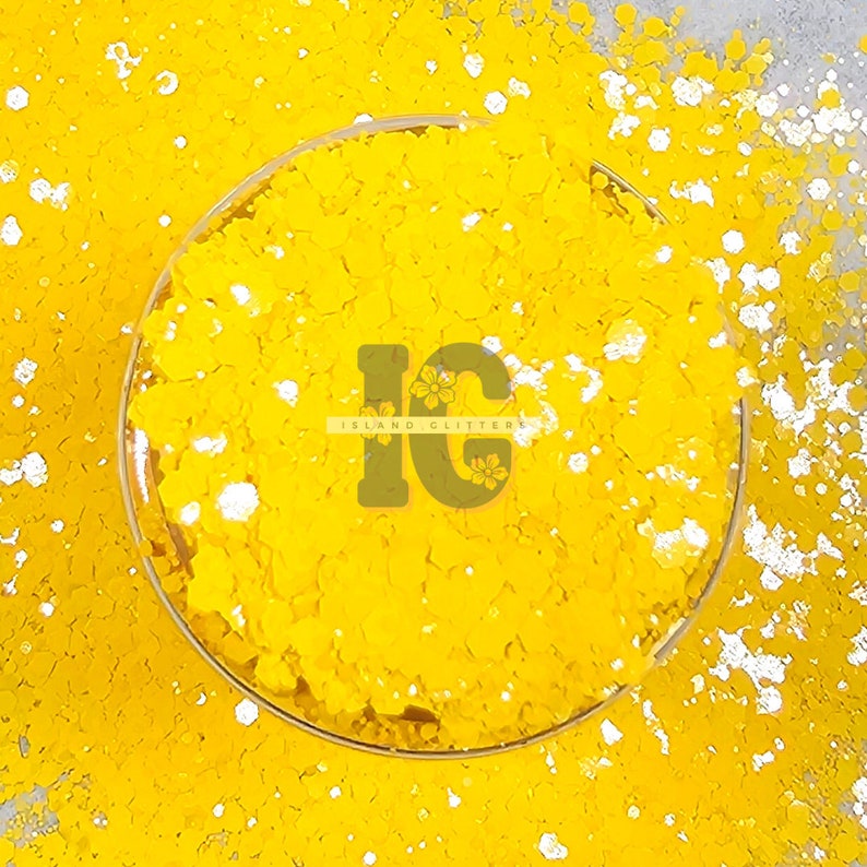 May include: A close-up of a yellow glitter with white glitter flakes. The glitter is in a clear glass jar with the text "Island Glitter" on the jar.