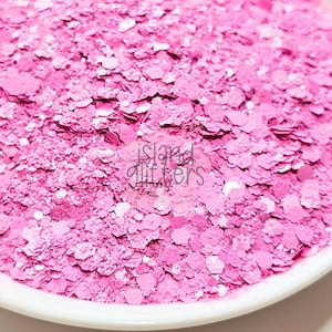 May include: Close-up of a pile of pink glitter with a mix of hexagonal and irregular shapes. The glitter is in a white bowl. The text "island glitters" is visible in the image.