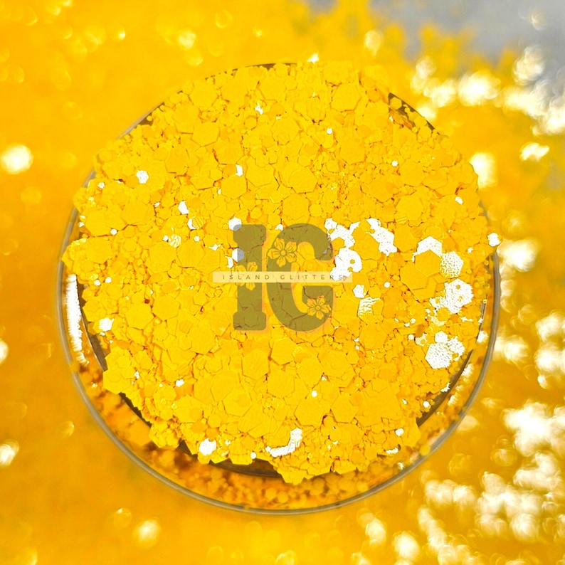 May include: Close-up of a yellow glitter, with a white iridescent sheen. The glitter is in a small container with the text "Island Glitter" and the letters "IG" in a stylized font.