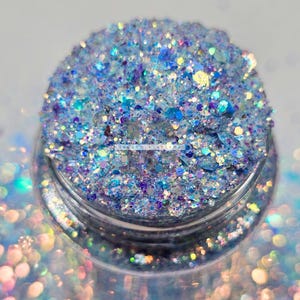 May include: A close-up of a jar filled with iridescent blue, purple, and white glitter. The glitter is a mix of different sizes and shapes, creating a sparkling effect.