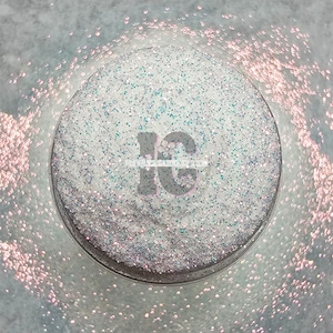 May include: A close-up of a white glitter with iridescent blue and pink flecks. The glitter is in a clear container.