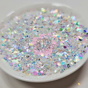 Puede incluir: Un plato de cerámica blanco lleno de una mezcla de purpurina iridiscente. La purpurina incluye varias formas y tamaños, reflejando un espectro de colores. El texto "island glitters" es visible en el centro del plato.