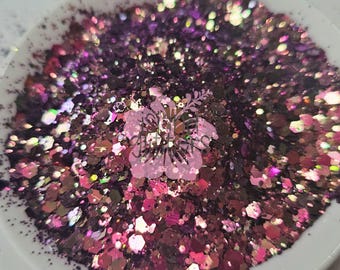 May include: A white bowl filled with a mix of glitter in shades of dark purple, pink, and iridescent colours. The glitter pieces are of various shapes and sizes, creating a sparkling effect. Ideal for craft projects.