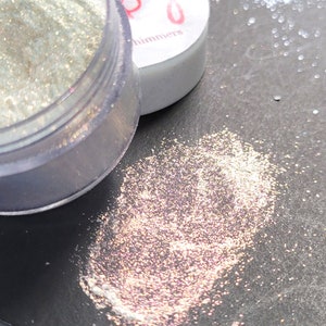 Puede incluir: Un pequeño frasco de purpurina iridiscente con una tapa blanca que dice "Shimmers". La purpurina es de color rosa claro y blanco y está esparcida sobre una superficie gris oscura.