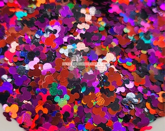 May include: A close-up of a bowl filled with iridescent holographic Mickey Mouse head shaped glitter. The glitter is a mix of red, purple, pink, blue, green, and black.