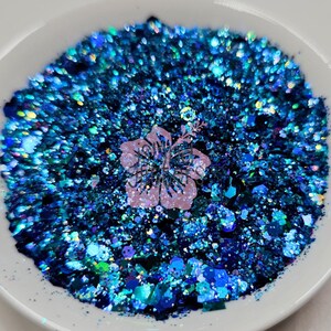 May include: A white bowl filled with a mix of blue and teal glitter. A light pink hibiscus flower is in the center of the glitter. The glitter is a mix of different shapes and sizes.