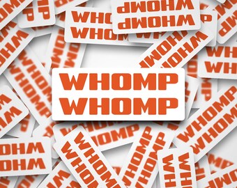 May include: A collection of white rectangular stickers with the word "WHOMP" in orange, scattered across a white background. The stickers are slightly angled, creating a sense of depth and movement. The text is bold and easy to read.