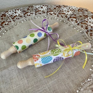 May include: Two wooden rolling pins decorated with colorful paper. One pin has a polka dot pattern in shades of green, pink, and yellow. The other pin has a pattern of colorful Easter eggs. Both pins are tied with ribbon.