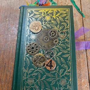 May include: A dark green hardcover book featuring a gold floral pattern on the cover. The front is decorated with metal clock gears, a circular wooden piece with the number 40, and a small wooden disc displaying the number 4.