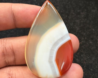 May include: A teardrop-shaped polished gemstone with a mix of colors. The stone features bands of white, orange, and clear sections. The gemstone is held in a hand, showcasing its smooth, reflective surface. This is a natural stone.