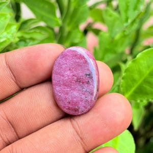 Pode incluir: Uma pedra preciosa cabochão de formato oval com uma paleta de cores magenta vibrante e rosa profundo, apresentando sutis inclusões minerais pretas por toda a superfície polida, com textura lisa e um acabamento reflexivo e brilhante.