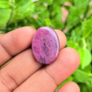 Può includere: Una gemma cabochon di forma ovale caratterizzata da una vibrante tavolozza di colori magenta e rosa intenso con inclusioni minerali nere sparse su tutta la superficie lucida, con una parte superiore liscia e arrotondata e finitura lucida.