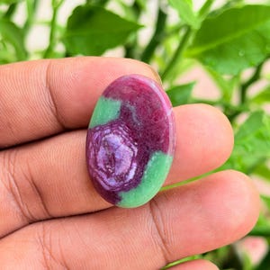 Pode incluir: Uma pedra preciosa polida de formato oval com uma combinação vibrante de cores vermelho magenta profundo e verde lima brilhante. A pedra possui uma superfície lisa e arredondada com inclusões minerais naturais visíveis e um acabamento brilhante.