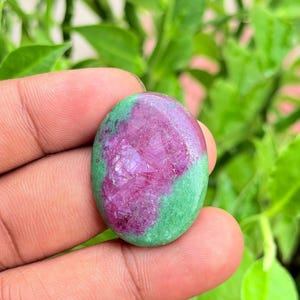 Op de afbeelding: Een ovaalvormige cabochon edelsteen met een levendig magenta-roze centrum omringd door een gevlekte groene rand. Het oppervlak van de steen vertoont een gepolijste, gladde textuur met zichtbare kristallijne structuren in het roze gedeelte.