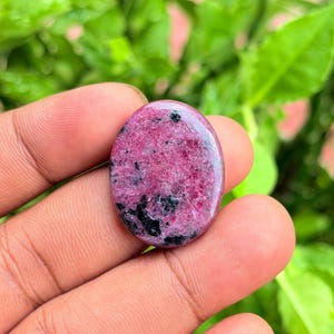 Può includere: Una gemma cabochon di forma ovale caratterizzata da un colore di base magenta e rosa intenso con inclusioni minerali nere irregolari sparse su tutta la superficie lucida, che presenta una consistenza liscia e brillante.