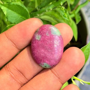 Op de afbeelding: Een ovaalvormige cabochon edelsteen met een levendig magenta-roze oppervlak met gevlekte patronen van lichtgroene minerale insluitingen door de hele steen, met een gladde, gepolijste en reflecterende textuur over het afgeronde oppervlak.