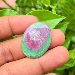 Puede incluir: Una piedra preciosa cabujón de forma ovalada con una superficie lisa y pulida, que presenta una mezcla vibrante de inclusiones minerales de color rosa magenta intenso y verde lima brillante, mostrando un patrón de color natural y veteado.