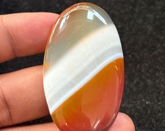 May include: Oval-shaped gemstone with a polished surface. The stone features bands of white, orange, and red-brown hues. The gemstone is held in a hand, showcasing its size and shape. The stone's colors and patterns are clearly visible.