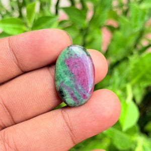 Puede incluir: Una piedra preciosa cabujón de forma ovalada con una superficie pulida que presenta un patrón de color distintivo de secciones rosa magenta vibrante que contrastan con áreas de color verde bosque profundo, con un acabado liso y brillante.