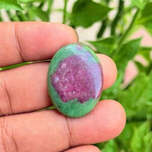 Può includere: Una gemma a cabochon di forma ovale caratterizzata da un vibrante centro magenta-rosa circondato da una matrice di zoisite verde opaca e liscia. La pietra presenta una superficie lucida e brillante che riflette la luce.