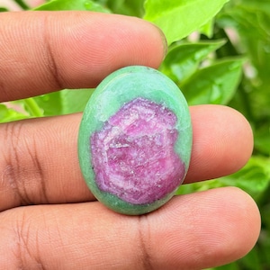 Può includere: Una gemma a cabochon di forma ovale caratterizzata da un centro rosa magenta vibrante circondato da un bordo liscio verde chiaro. La pietra presenta una superficie lucida e brillante con una netta distinzione cromatica tra il nucleo rosa e lo strato verde.