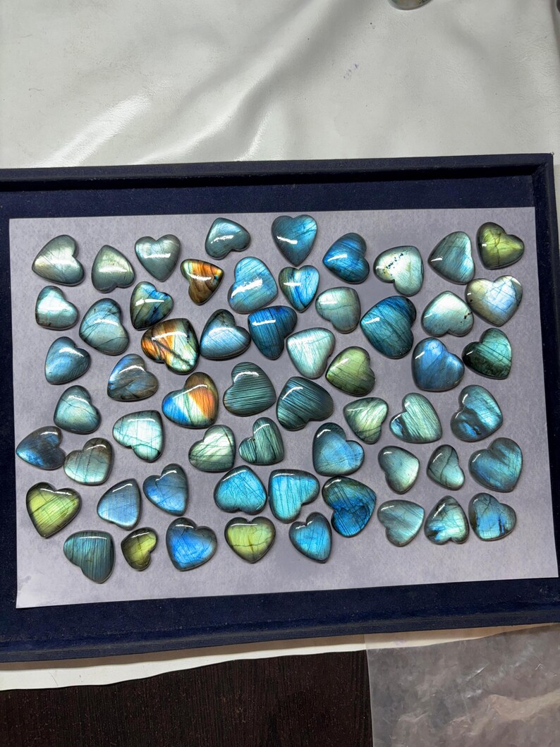 May include: A collection of heart-shaped labradorite gemstones in various shades of blue, green, and gold. The stones are arranged in a display box.
