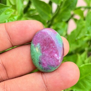 Puede incluir: Una piedra preciosa pulida de forma ovalada que presenta una mezcla vibrante de colores magenta rosado intenso y verde bosque. La piedra tiene una superficie lisa y brillante con texturas cristalinas naturales visibles.