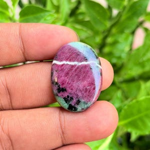 May include: An oval-shaped polished ruby zoisite gemstone featuring a vibrant magenta-pink centre, patches of deep green, and scattered black mineral inclusions, accented by a distinct horizontal white quartz vein running across the surface.