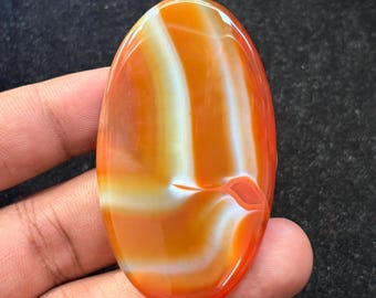 May include: Oval-shaped polished gemstone with a vibrant orange and white banded pattern. The stone has a glossy finish and is held in a hand, showcasing its natural beauty. The gemstone's design is reminiscent of agate.