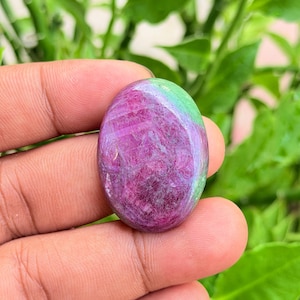 Puede incluir: Una piedra preciosa cabujón de forma ovalada que presenta un centro prominente de color magenta y púrpura intenso con una sección verde vibrante distintiva en la parte superior. La piedra tiene una superficie lisa y pulida.