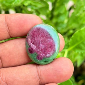 Puede incluir: Una piedra preciosa pulida de forma ovalada que presenta un centro de color rosa magenta vibrante rodeado por un borde verde claro liso y moteado. La piedra tiene una superficie brillante y reflectante con texturas cristalinas naturales.