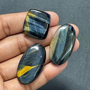 May include: Three polished gemstones featuring a chatoyant blue and gold tiger eye pattern. One stone is square-shaped with rounded corners, while the other two are oval-shaped. The stones exhibit a smooth, reflective surface with distinct bands.