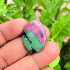 Pode incluir: Uma pedra preciosa polida de formato oval com uma divisão de cores distinta, apresentando uma seção rosa magenta vibrante e uma seção verde brilhante, acentuada por pequenas inclusões minerais pretas na superfície lisa e lustrosa.