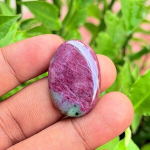 Pode incluir: Uma pedra preciosa cabochão de formato oval com uma superfície polida em tons de magenta e rosa profundo, acentuada por uma pequena mancha de inclusões minerais verde-escuras na base, apresentando uma textura lisa, arredondada e reflexiva.