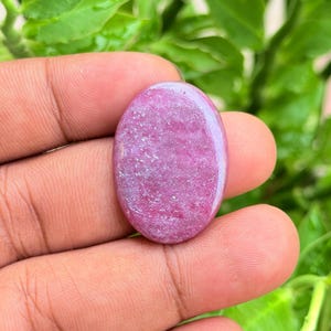 Puede incluir: Una piedra preciosa pulida de forma ovalada con un vibrante color rosa magenta y una superficie lisa y reflectante. La piedra presenta una textura granular sutil en todo su cuerpo, mostrando variaciones naturales en su tono rosa.