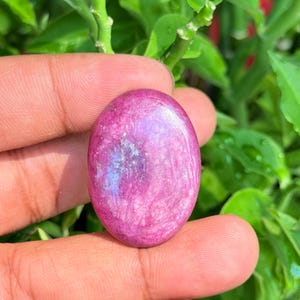 Peut inclure: Une pierre précieuse cabochon de forme ovale à la surface polie, présentant une palette de couleurs magenta vif et rose profond avec de subtiles inclusions irisées bleues et blanches visibles dans la structure cristalline translucide.