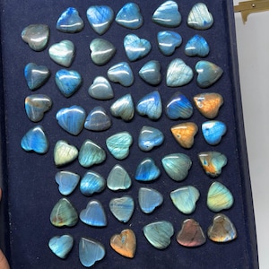 May include: A collection of 40 heart-shaped labradorite stones in various shades of blue, green, and gold. The stones are arranged in a display box with a dark blue velvet lining.