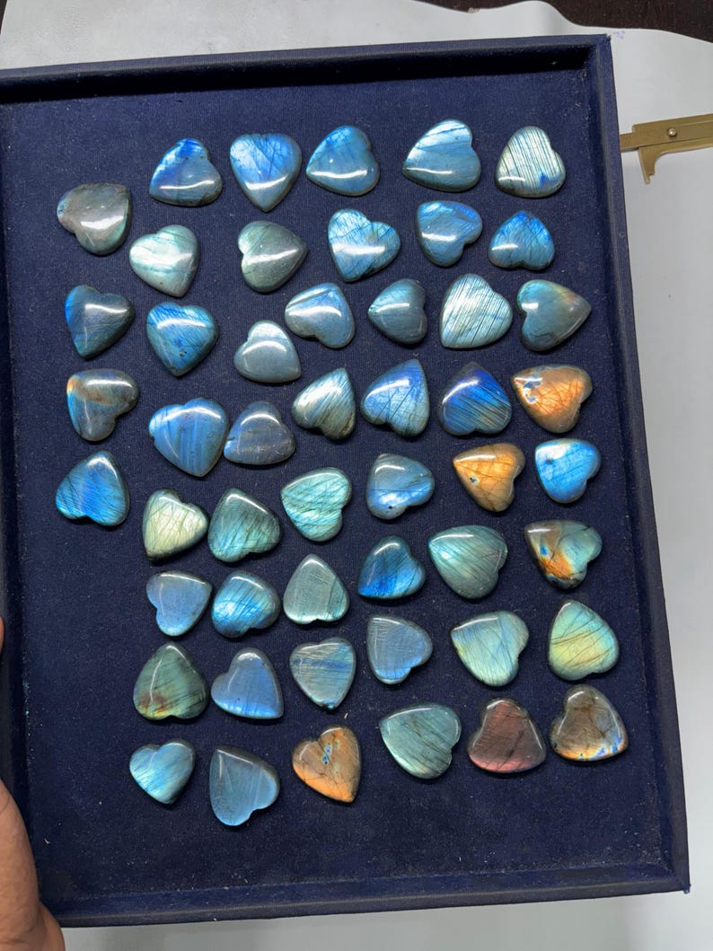 May include: A collection of 40 heart-shaped labradorite stones in various shades of blue, green, and gold. The stones are arranged in a display box with a dark blue velvet lining.