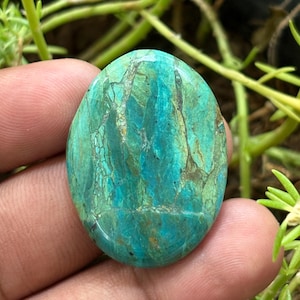 Könnte beinhalten: Ovaler Edelstein in Türkis-, Grün- und Blautönen. Der polierte Stein zeigt ein natürliches, marmoriertes Muster. Der Edelstein wird in einer Hand gehalten, mit grünem Laub im Hintergrund.