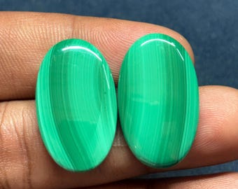 May include: Two oval-shaped malachite gemstones. The gemstones are a vibrant green color with distinctive banded patterns. The polished stones are smooth and reflect light, showcasing the natural variations in the mineral's structure. The gemstones are held in a person's hand.