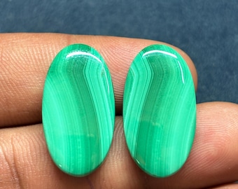 May include: Two polished oval malachite gemstones. The gemstones are a vibrant green color with darker green, banded patterns. The stones are smooth and have a glossy finish, reflecting light. The gemstones are held in a hand.