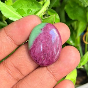 Puede incluir: Una piedra preciosa pulida de forma ovalada que presenta una clara división de color entre una sección rosa magenta vibrante y una sección verde menta suave. La piedra tiene una superficie lisa y brillante con inclusiones minerales.