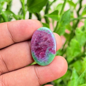 Pode incluir: Uma pedra preciosa em formato de cabochão oval com uma superfície polida, apresentando uma mistura vibrante de inclusões minerais em vermelho magenta profundo e verde brilhante. A pedra exibe um topo liso e arredondado com contraste.