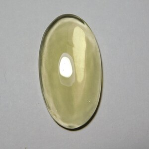 May include: A translucent, oval-shaped gemstone with a pale yellow hue. The polished surface reflects light, creating a bright spot in the center. The gemstone is set against a neutral background.