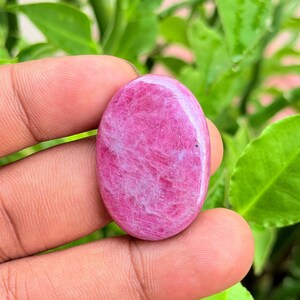 Pode incluir: Uma pedra preciosa polida de formato oval com uma paleta de cores magenta e rosa vibrantes. A pedra apresenta uma superfície lisa e brilhante com texturas fibrosas internas visíveis e inclusões brancas sutis na estrutura.